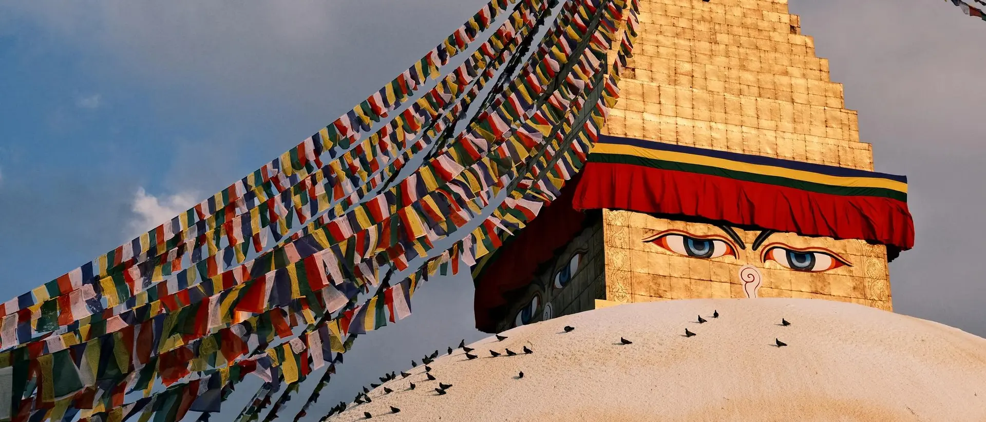 Boudhanath Stupa, adorned with colorful prayer flags, stands under a cloudy sky, with an airplane flying overhead.