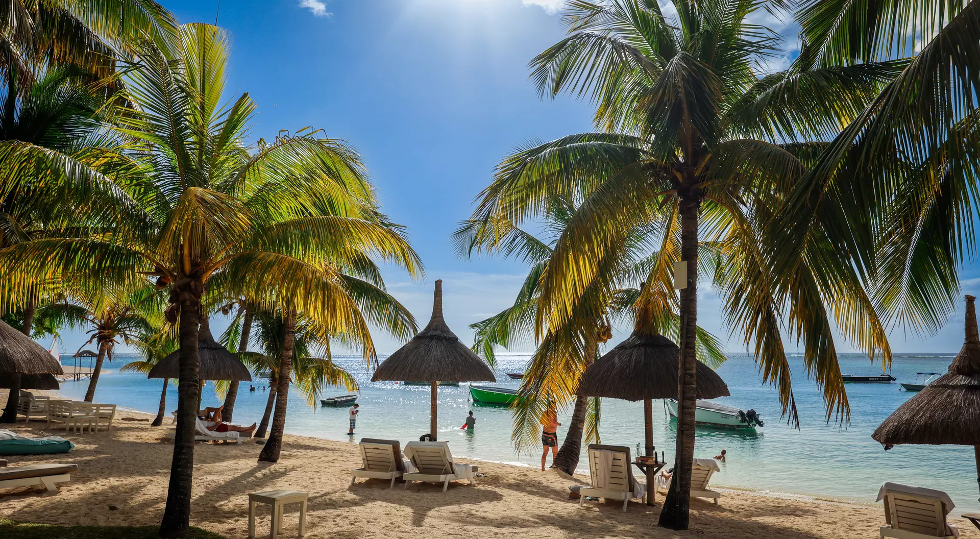 Tropical beach scene with palm trees, thatched umbrellas, and people enjoying the sunny day by the water.
