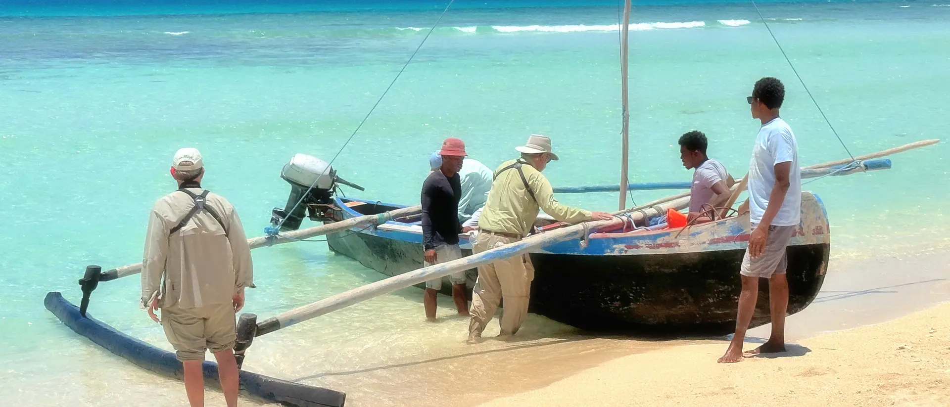 Five people working together to unload a boat on a sandy beach with clear turquoise water in the background.