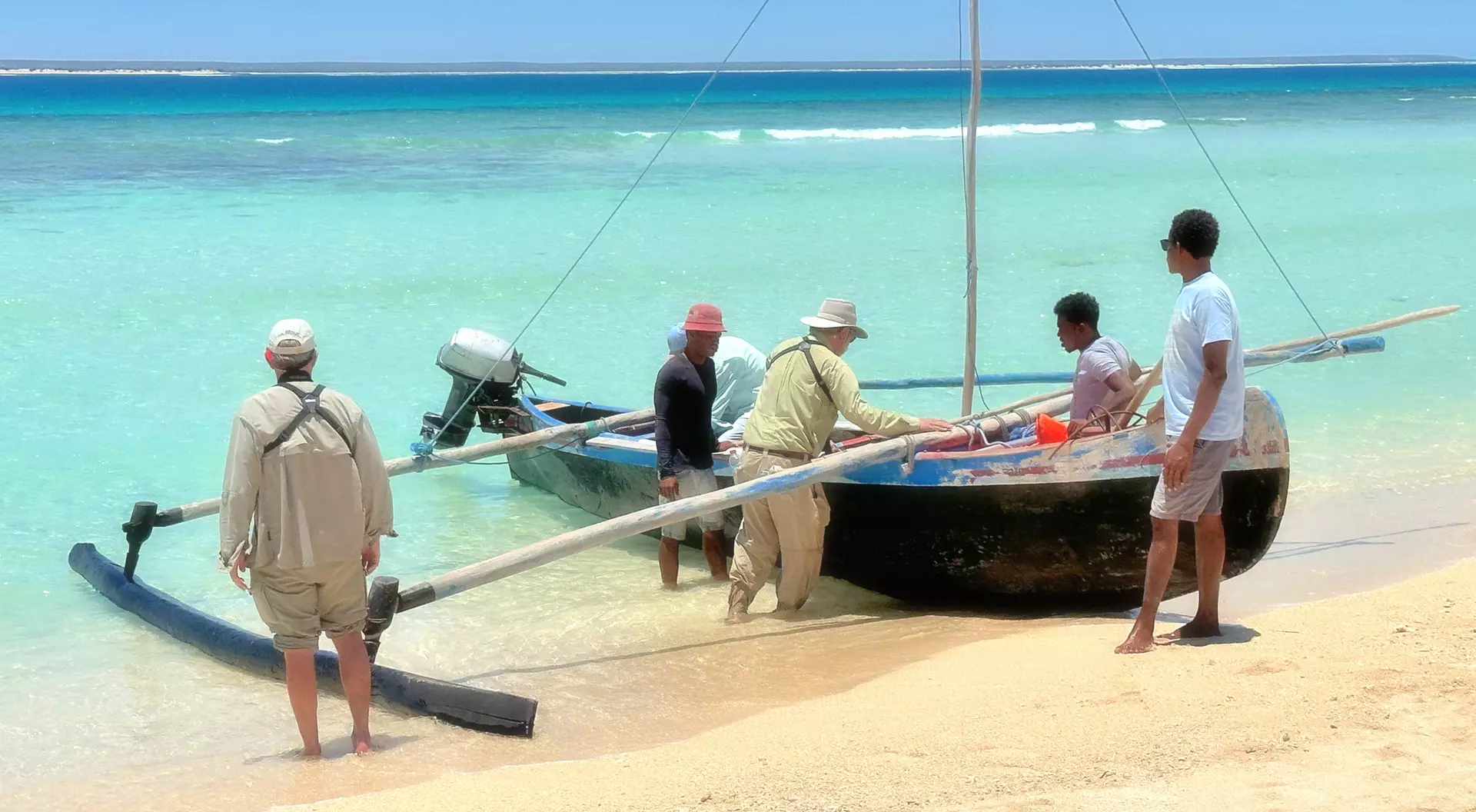Five people working together to unload a boat on a sandy beach with clear turquoise water in the background.