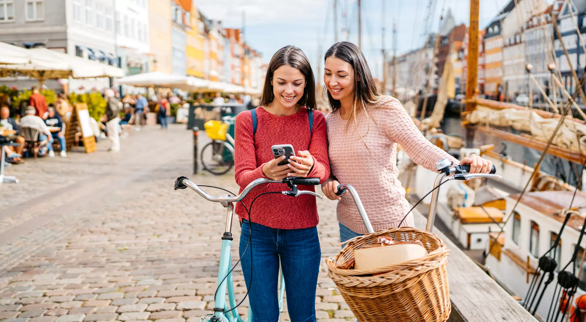 Two women smile while looking at a phone, with a bicycle and scenic harbor in the background.