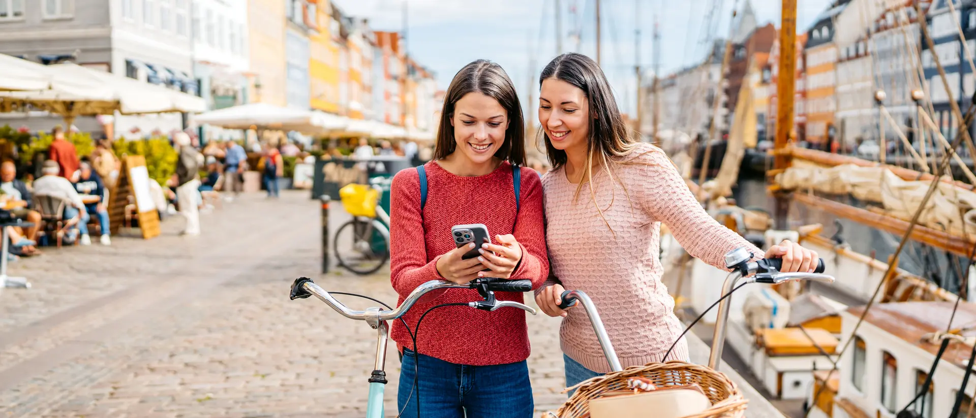 Two women smile while looking at a phone, with a bicycle and scenic harbor in the background.