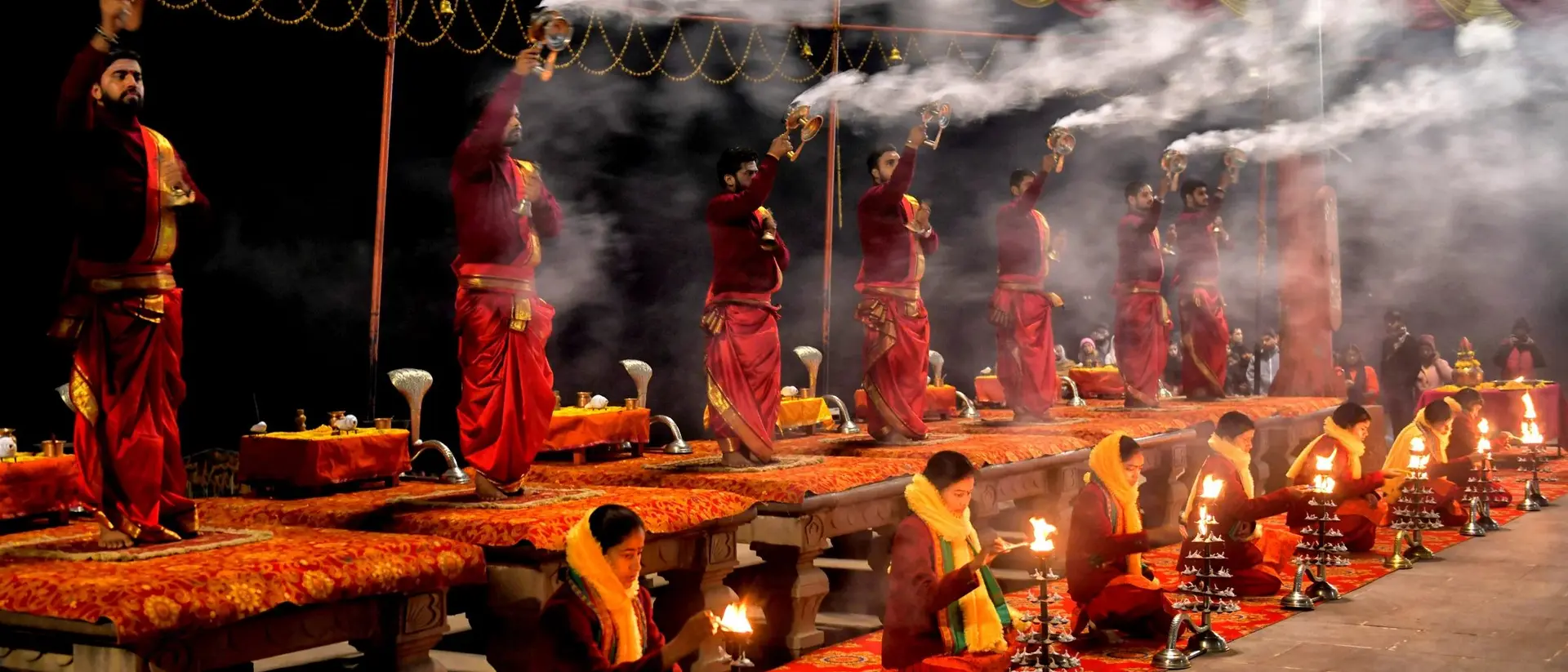 A group of individuals in traditional attire perform a ritual with lamps and incense, surrounded by smoke and bright decorations.