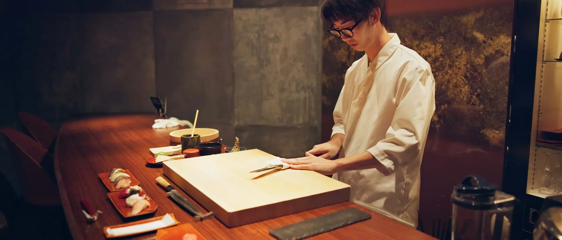 A sushi chef in a white uniform skillfully prepares sushi at a wooden counter, surrounded by sushi plates and tools.