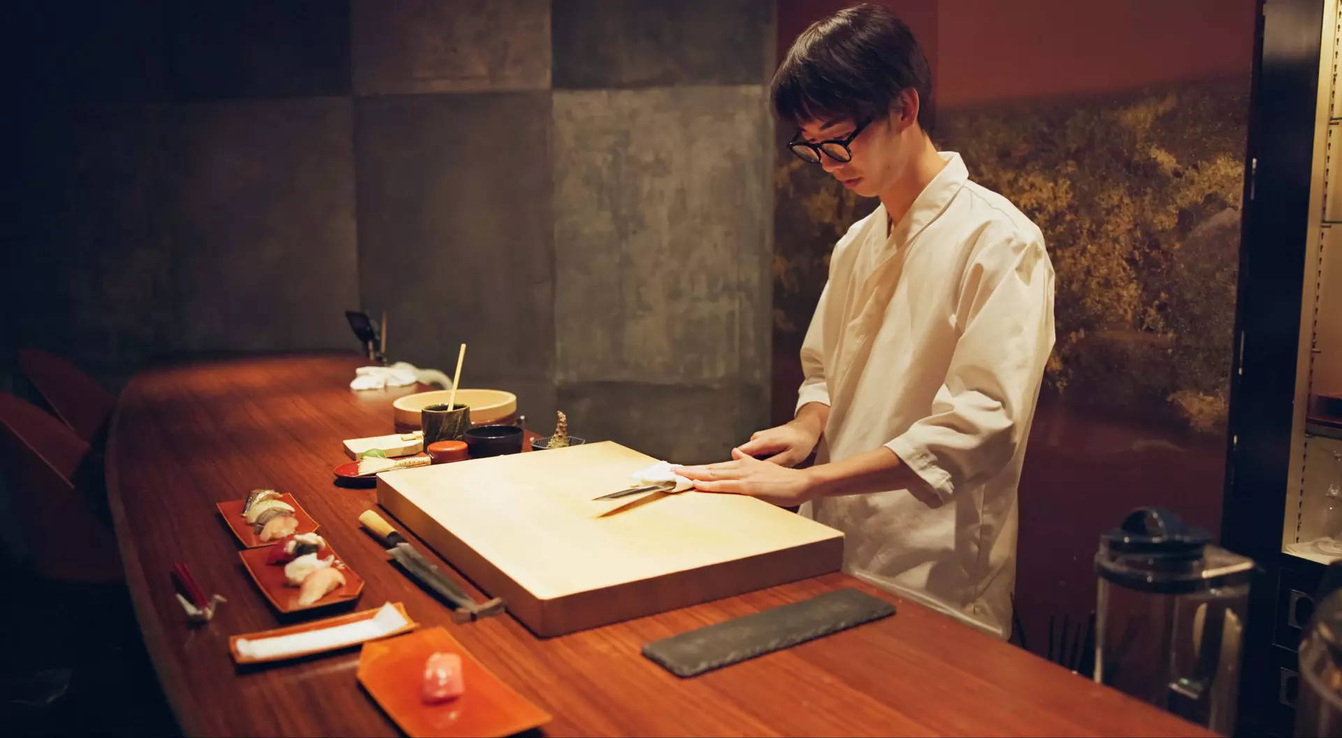 A sushi chef in a white uniform skillfully prepares sushi at a wooden counter, surrounded by sushi plates and tools.