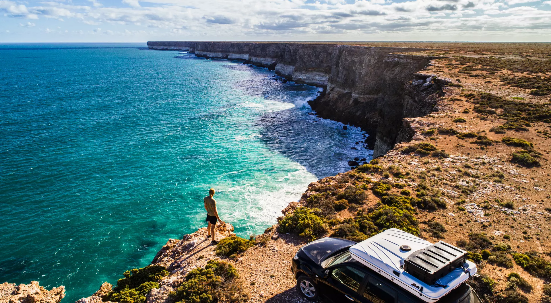 A person stands on a cliff edge overlooking a turquoise ocean, with a parked car nearby and scenic rock formations in view.