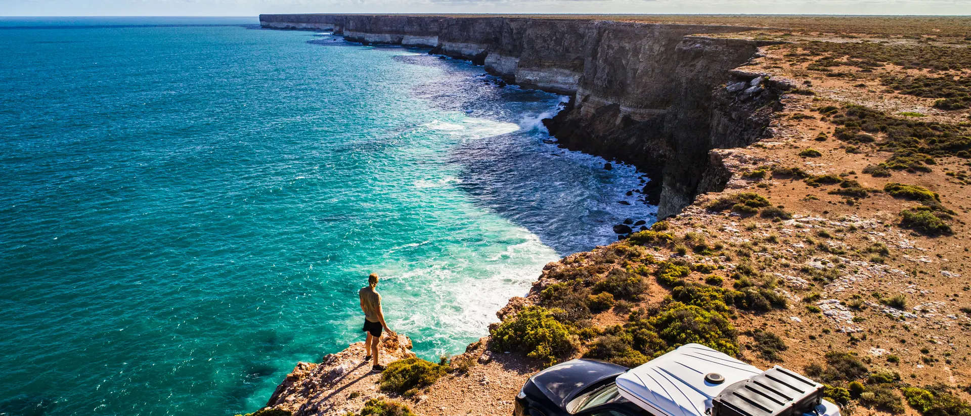 A person stands on a cliff edge overlooking a turquoise ocean, with a parked car nearby and scenic rock formations in view.