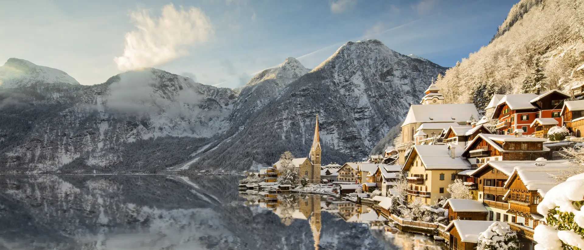 Snow-covered village by a lake, surrounded by mountains, with a church steeple reflecting in the water.