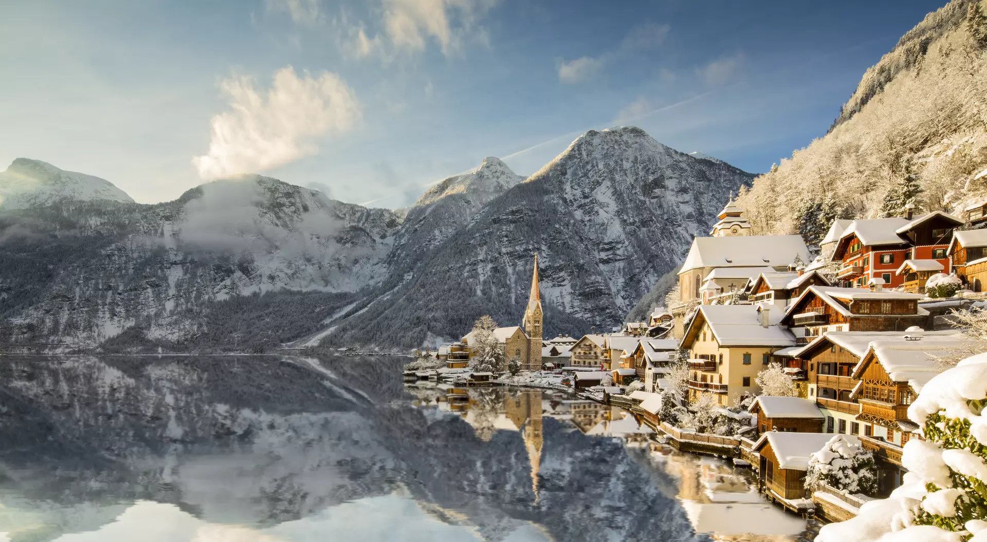 Snow-covered village by a lake, surrounded by mountains, with a church steeple reflecting in the water.