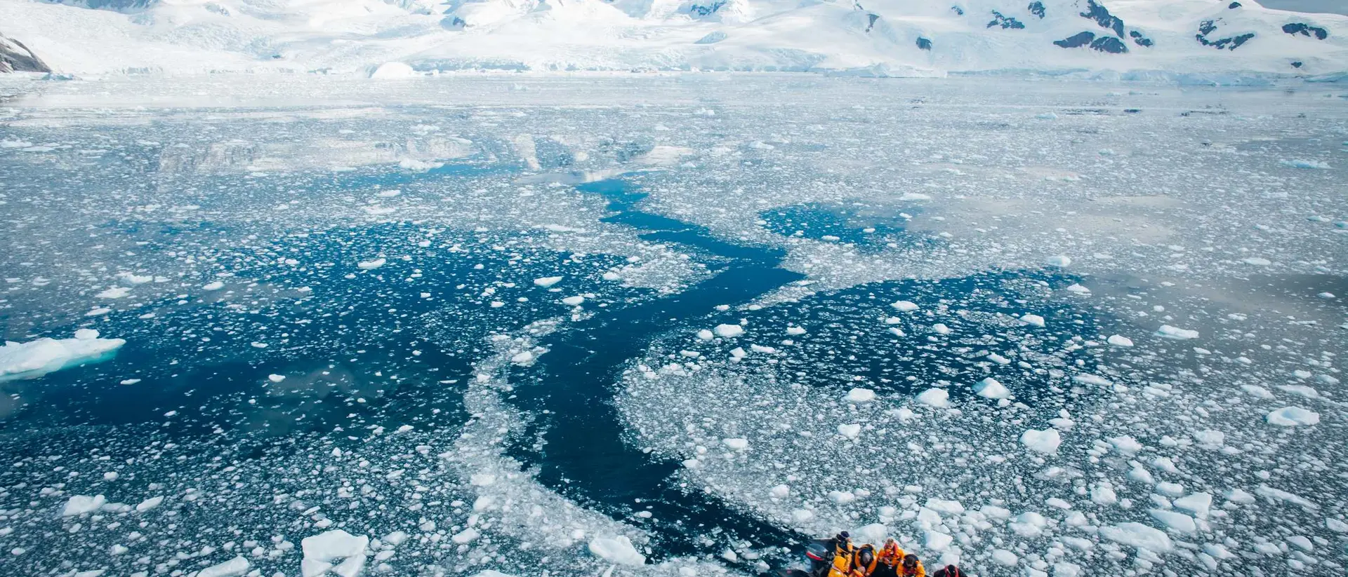 A small boat navigates through icy waters with snow-capped mountains in the background.