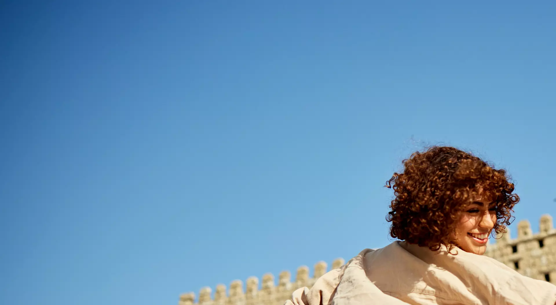 Young woman with curly hair joyfully walking near a stone archway, with a clear blue sky in the background.