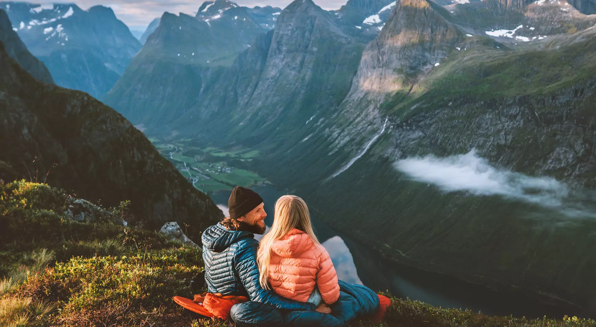 A couple sits together on a grassy hillside, enjoying scenic mountain views during sunset.