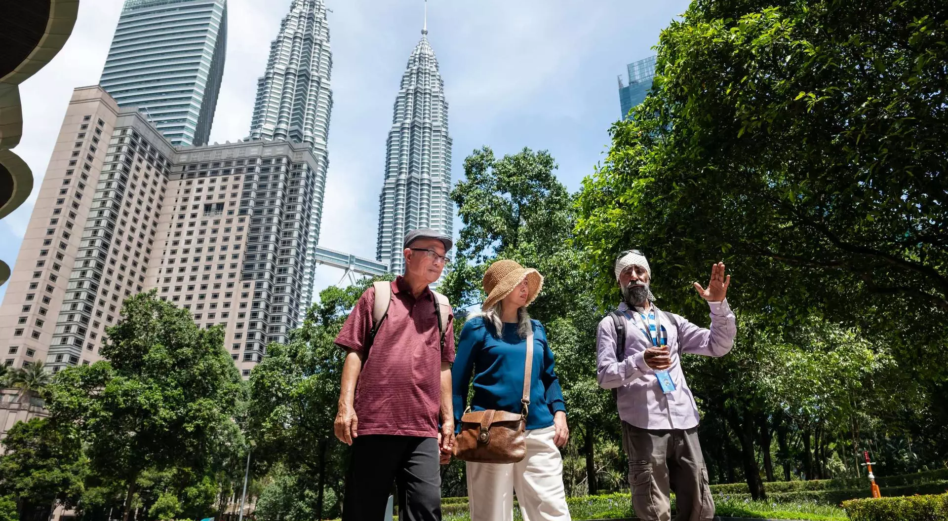 Three people walk together in a park with tall buildings visible in the background under a clear blue sky.