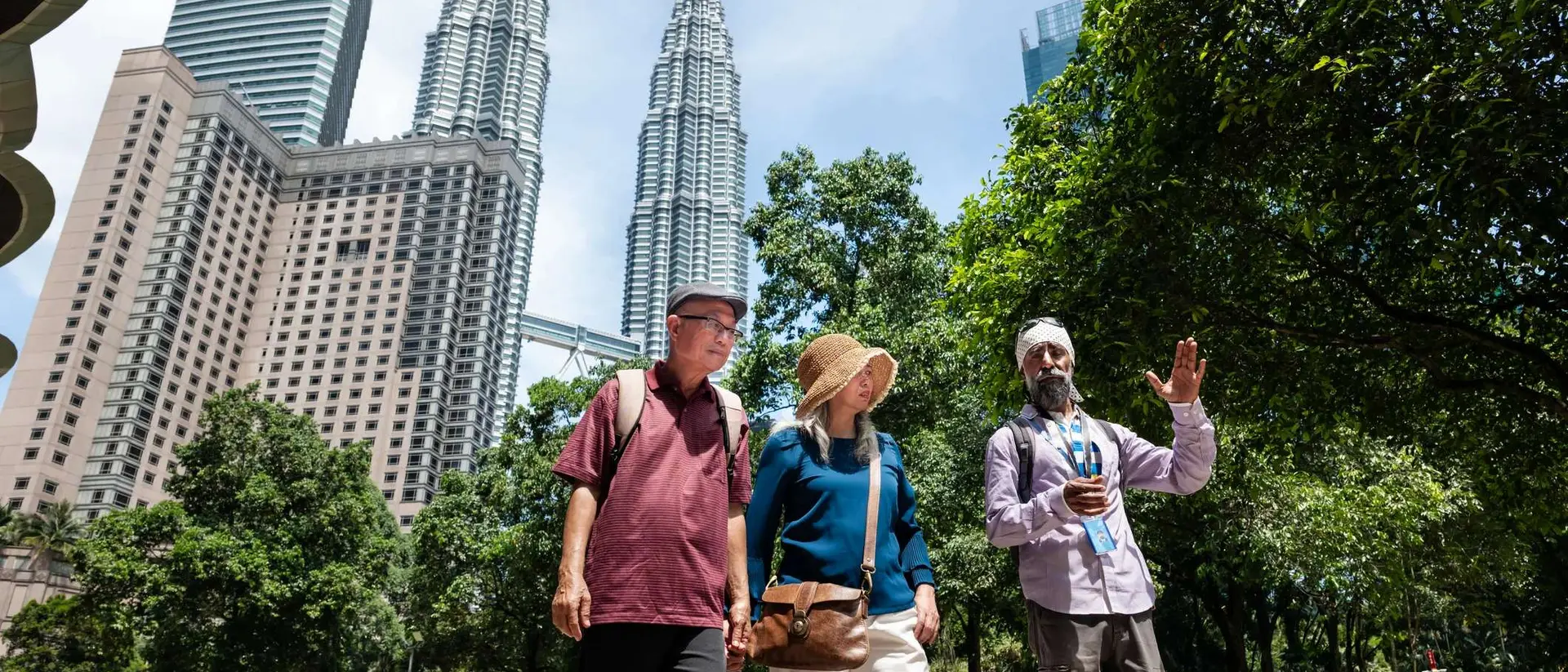 Three people walk together in a park with tall buildings visible in the background under a clear blue sky.