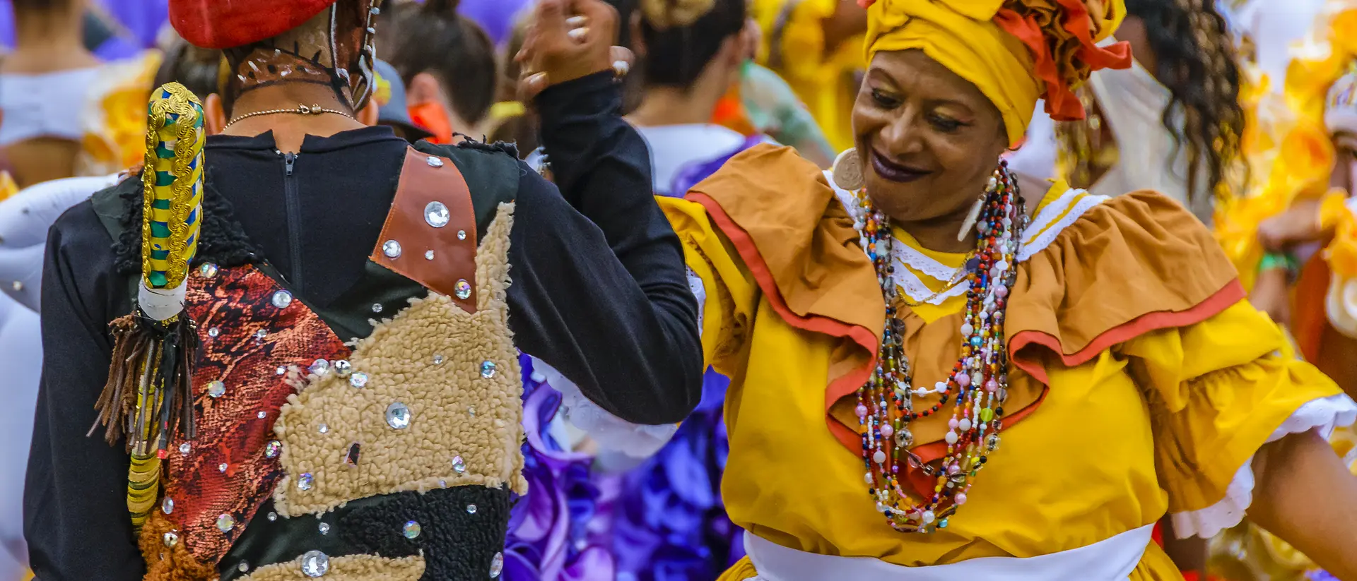 Two dancers in colorful traditional outfits engage in a joyful dance, surrounded by a festive crowd.