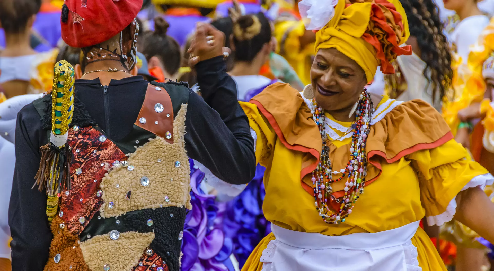 Two dancers in colorful traditional outfits engage in a joyful dance, surrounded by a festive crowd.