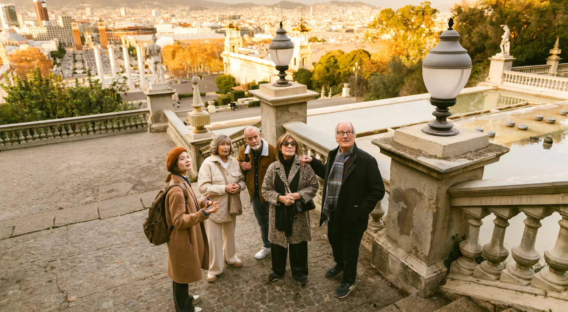 A group of five people stands on a staircase overlooking a cityscape at sunset. Trees and decorative lampposts nearby.