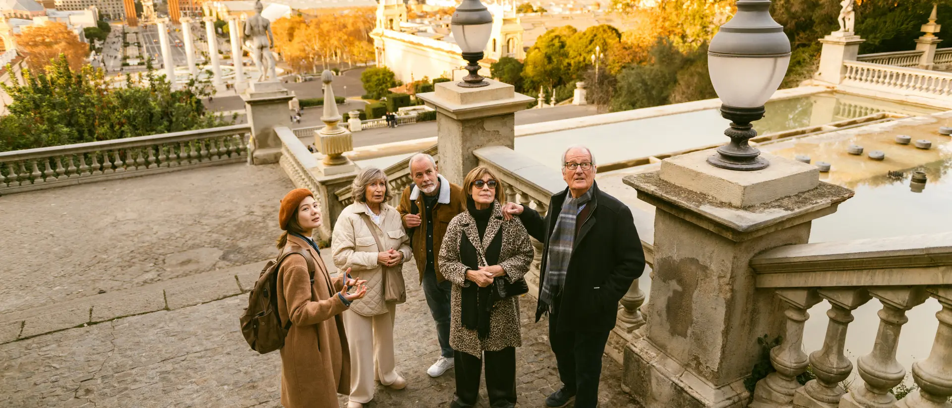 A group of five people stands on a staircase overlooking a cityscape at sunset. Trees and decorative lampposts nearby.