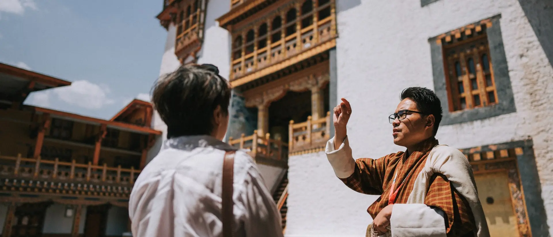 A tour guide gestures while speaking to a visitor outside a traditional Bhutanese building on a sunny day.