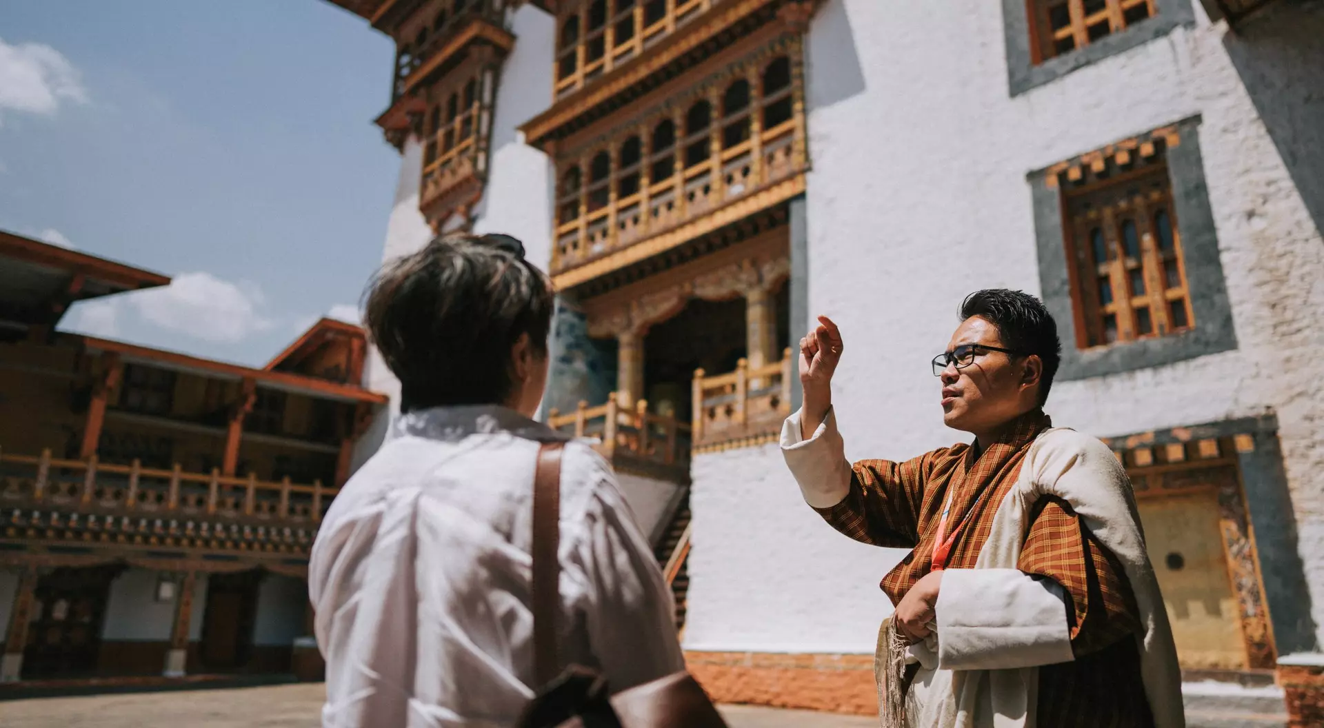 A tour guide gestures while speaking to a visitor outside a traditional Bhutanese building on a sunny day.