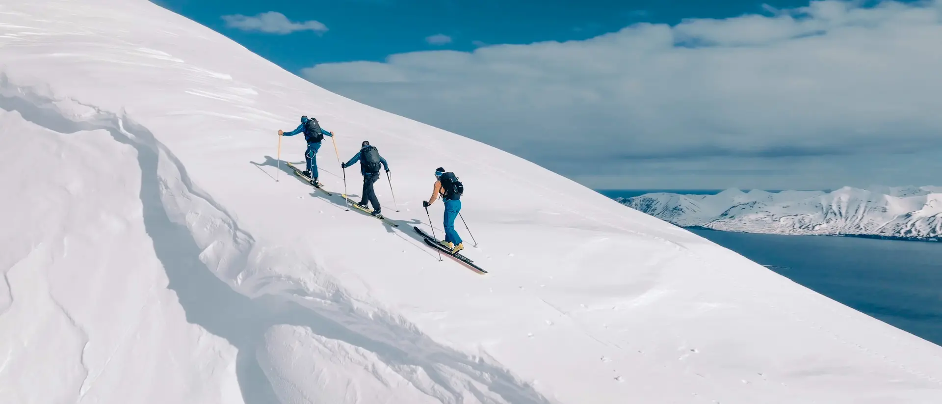 Three skiers ascend a snowy slope under a bright blue sky, with mountains and water visible in the background.