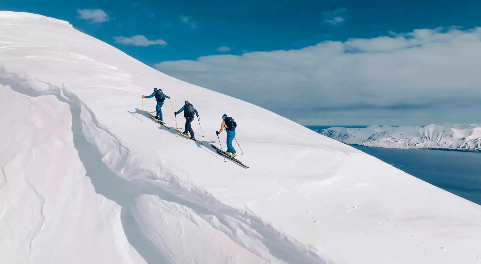 Three skiers ascend a snowy slope under a bright blue sky, with mountains and water visible in the background.