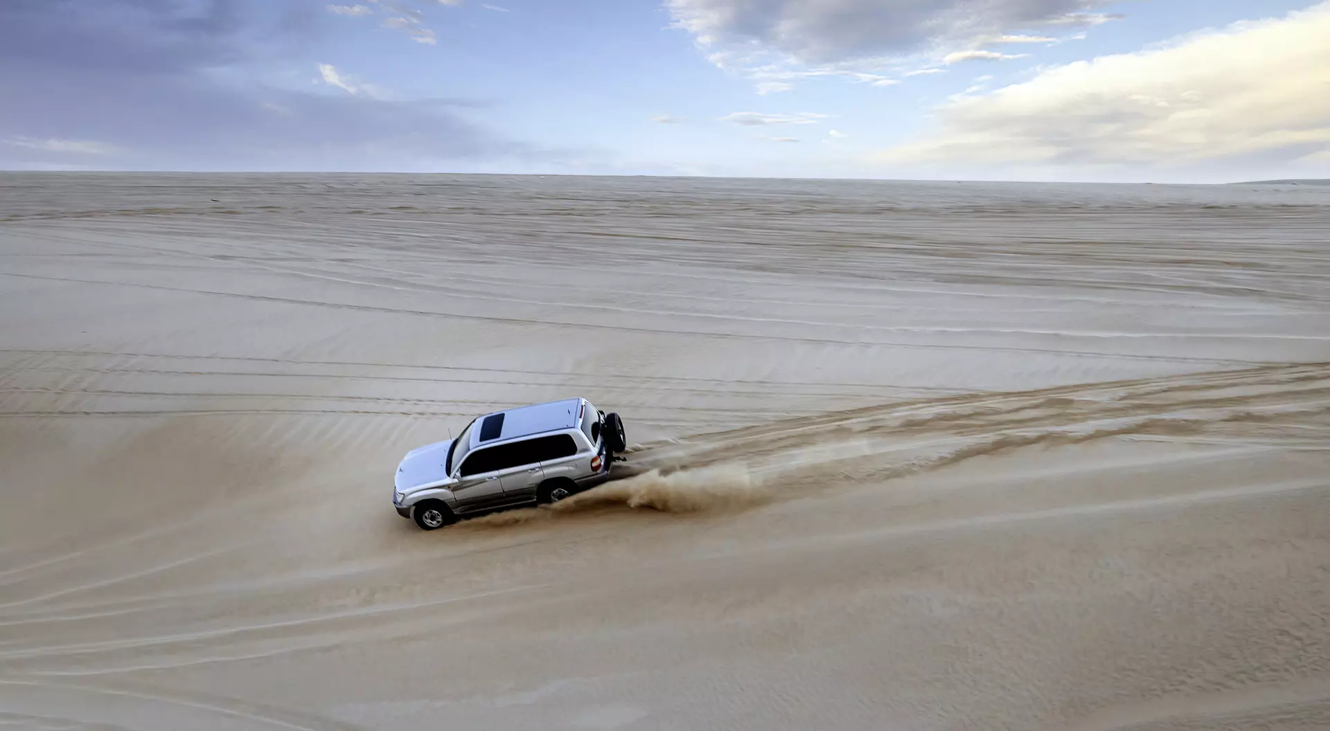 A white SUV drives over sandy dunes under a cloudy sky, leaving tire tracks in the soft sand.