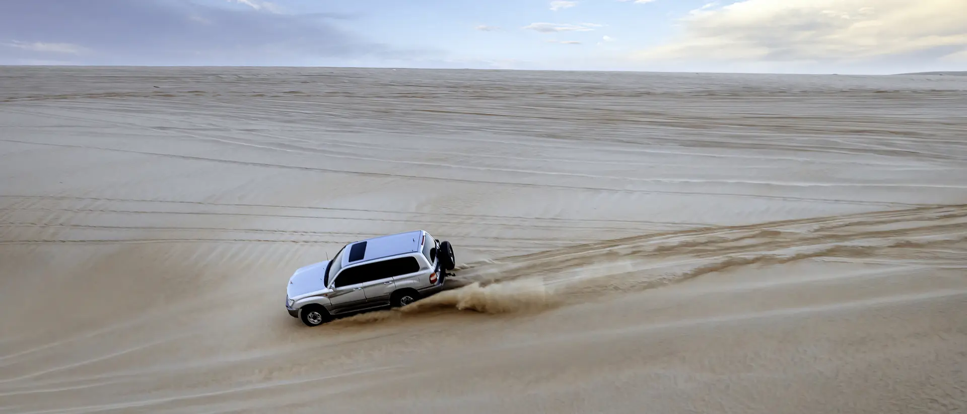 A white SUV drives over sandy dunes under a cloudy sky, leaving tire tracks in the soft sand.