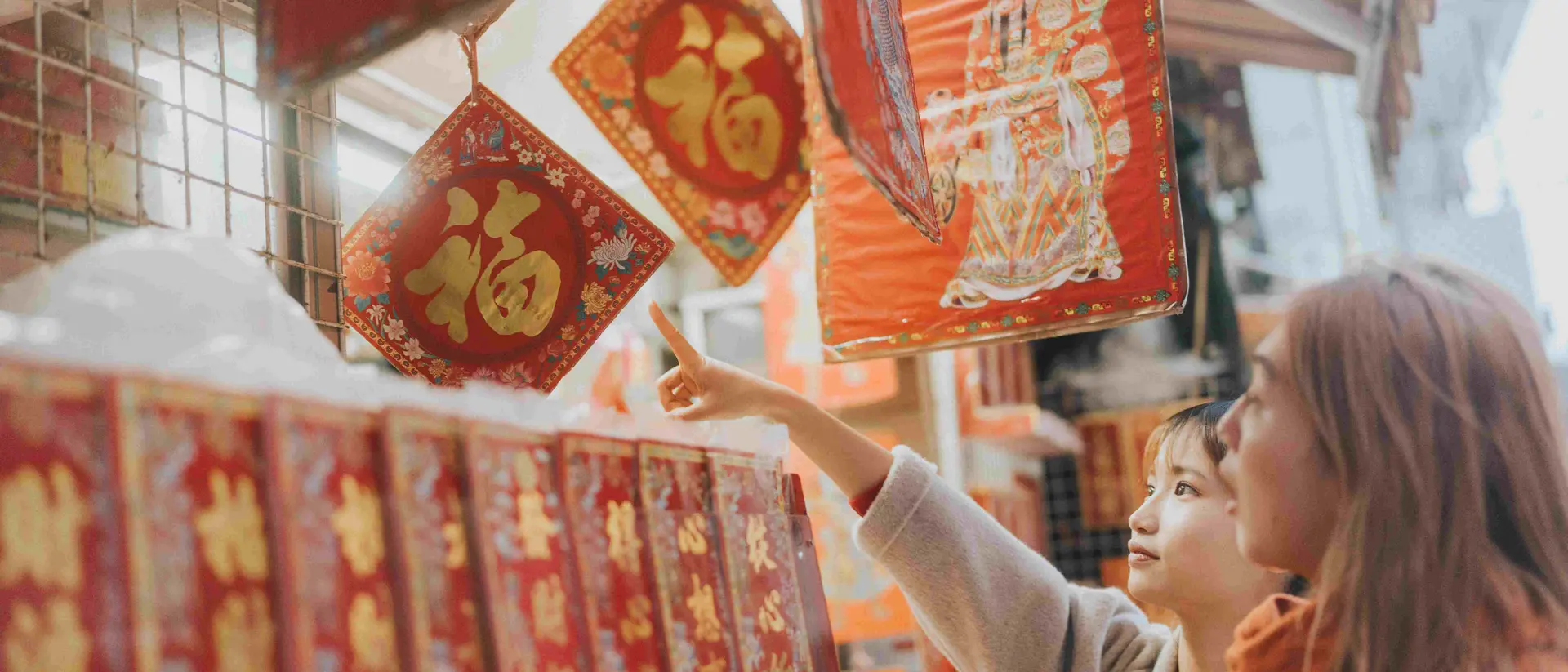 Two women admire decorative red and gold items hanging at a market, pointing at one while browsing the scene.
