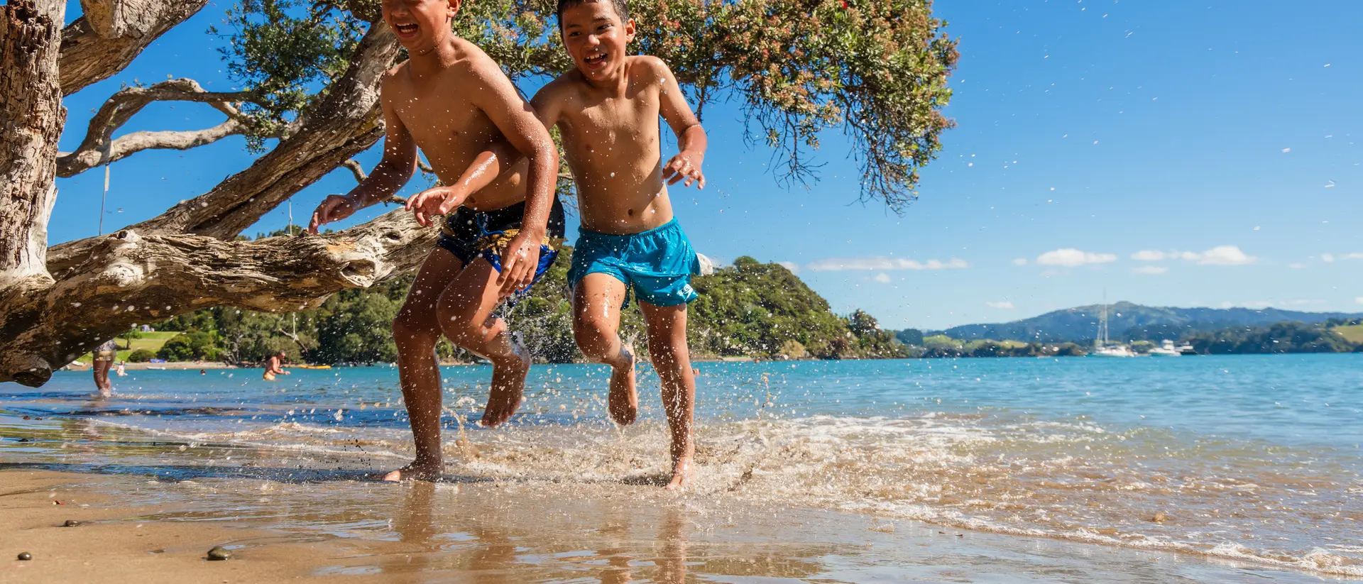 Two boys run joyfully through shallow water on a sunny beach, with trees and hills in the background.