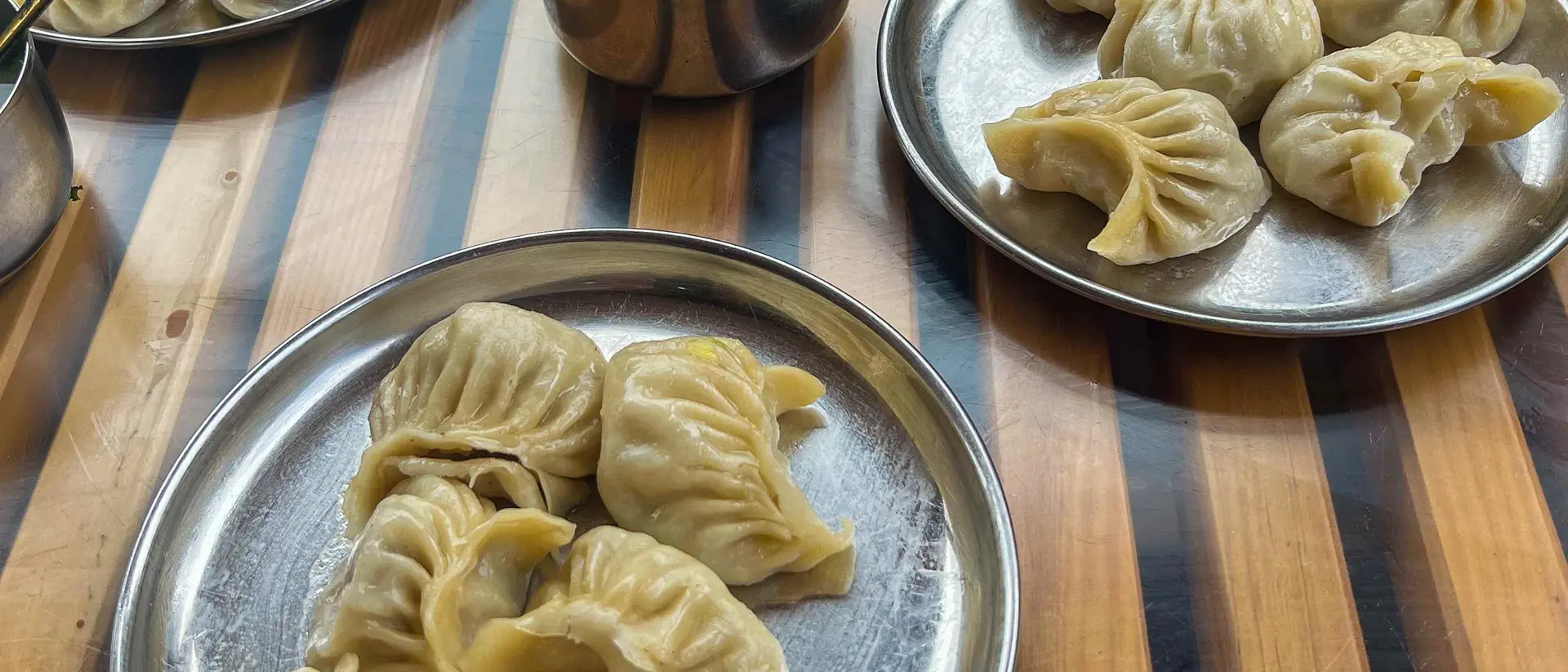Three metal plates hold steamed dumplings, with a bowl of sauce on a wooden table.