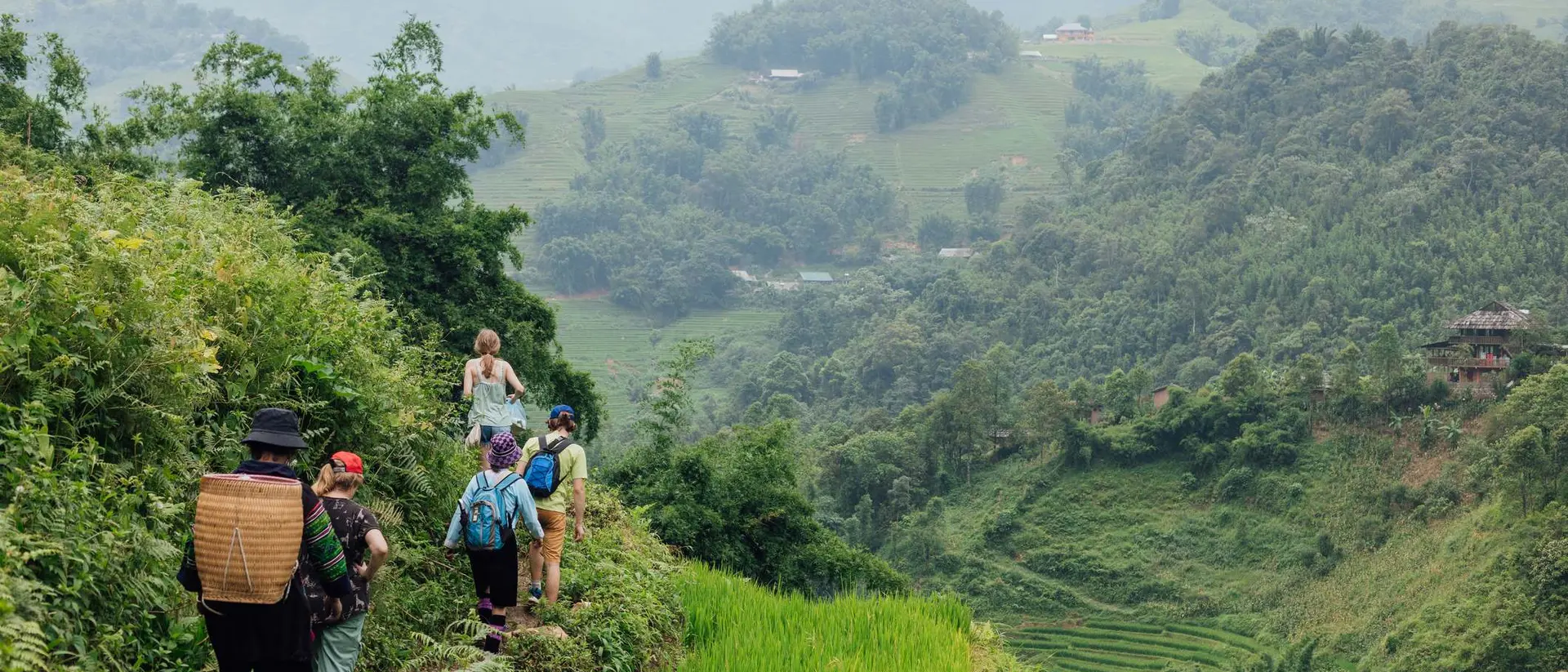Group of hikers walking along a narrow path through lush green rice terraces with mountains in the background.