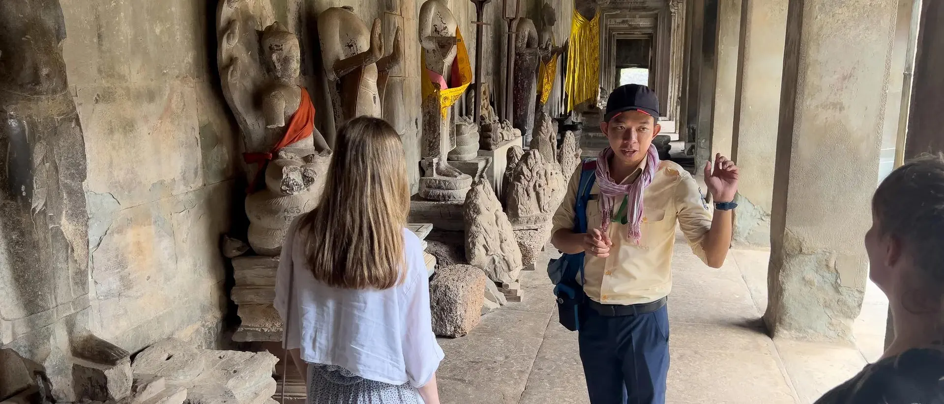 Tour guide talking to two visitors in a historic corridor with Buddha statues and decorative cloth in the background.