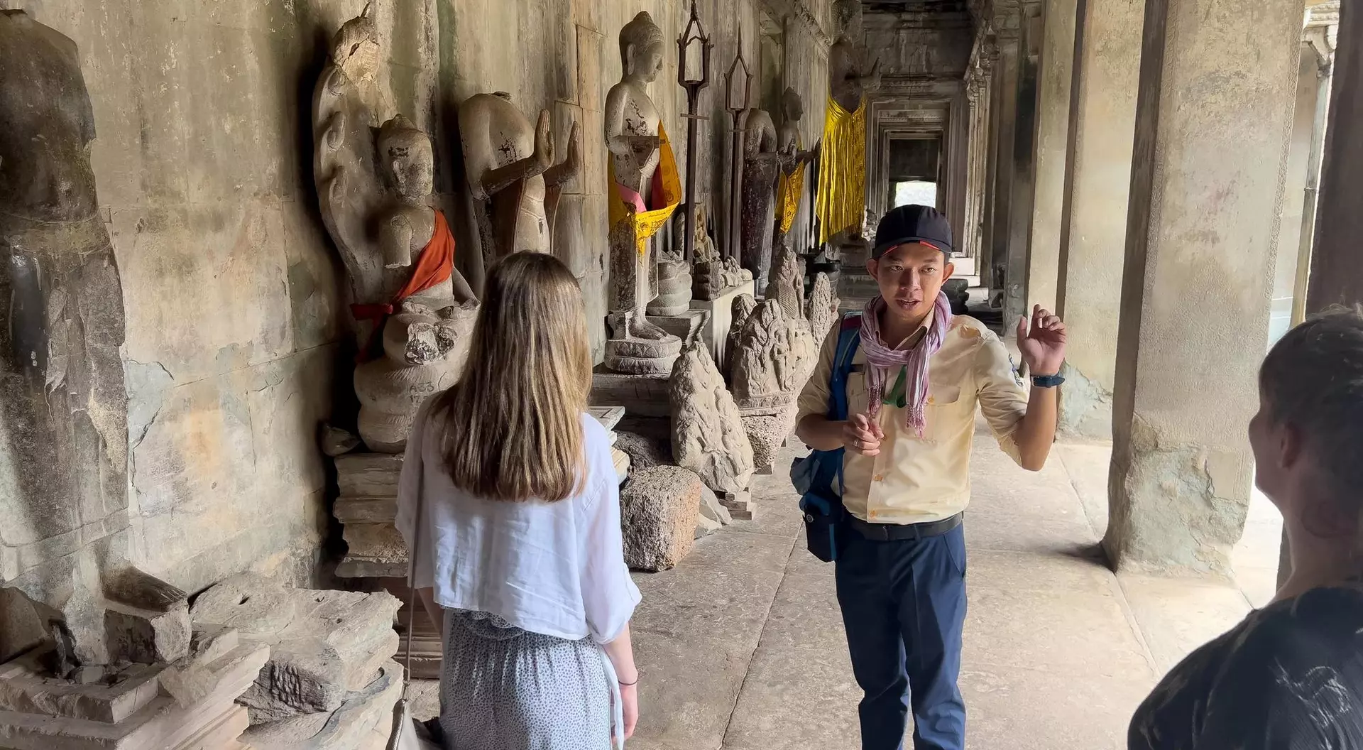 Tour guide talking to two visitors in a historic corridor with Buddha statues and decorative cloth in the background.