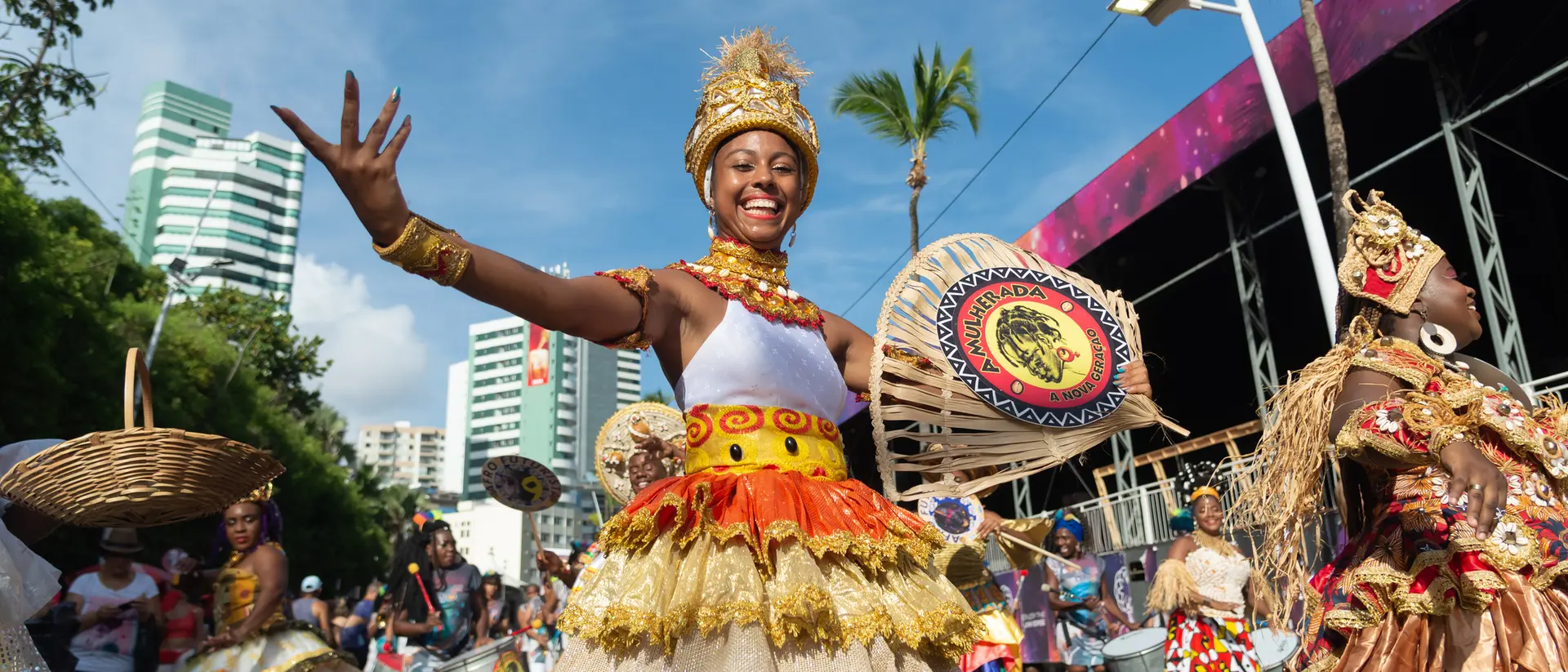 A smiling dancer in vibrant costume celebrates during a lively parade, surrounded by colorful decorations and onlookers.