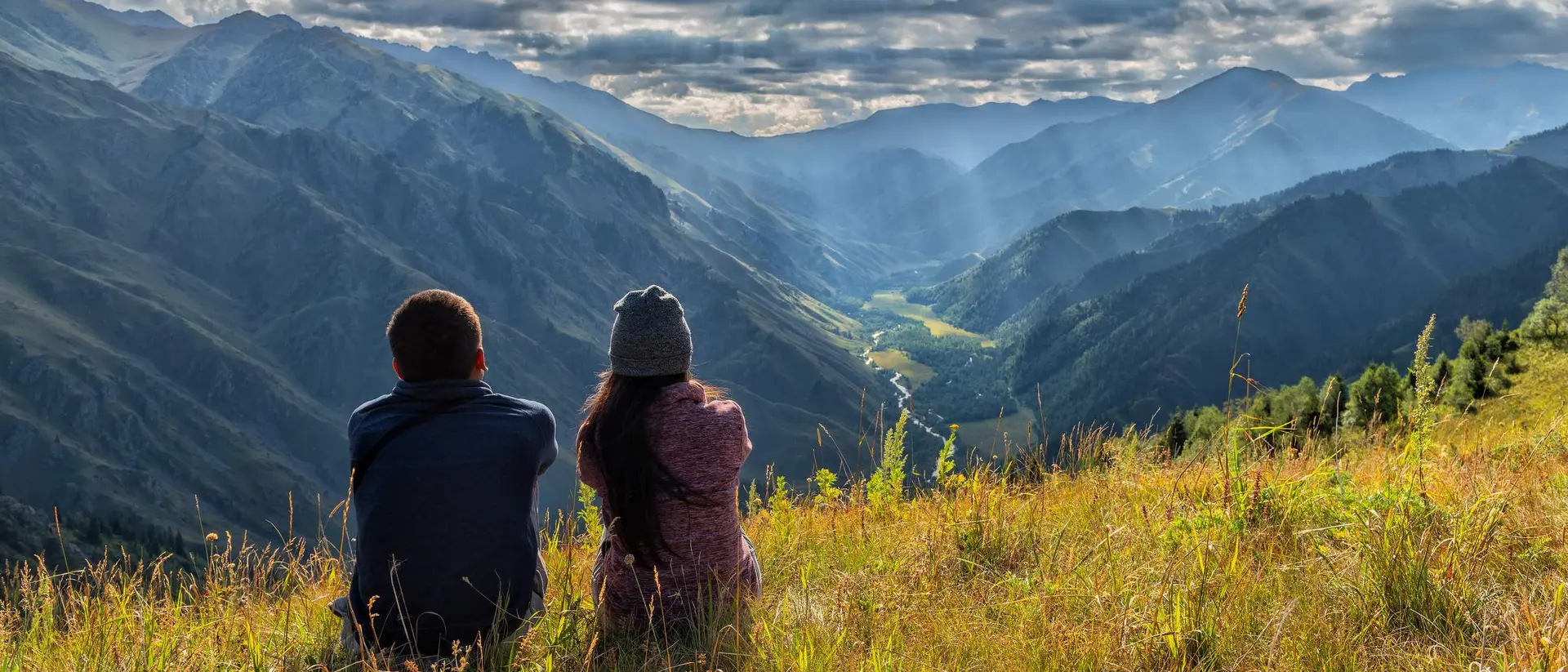 Two people sit on a grassy hillside, gazing at a panoramic view of mountains and valleys under a cloudy sky.