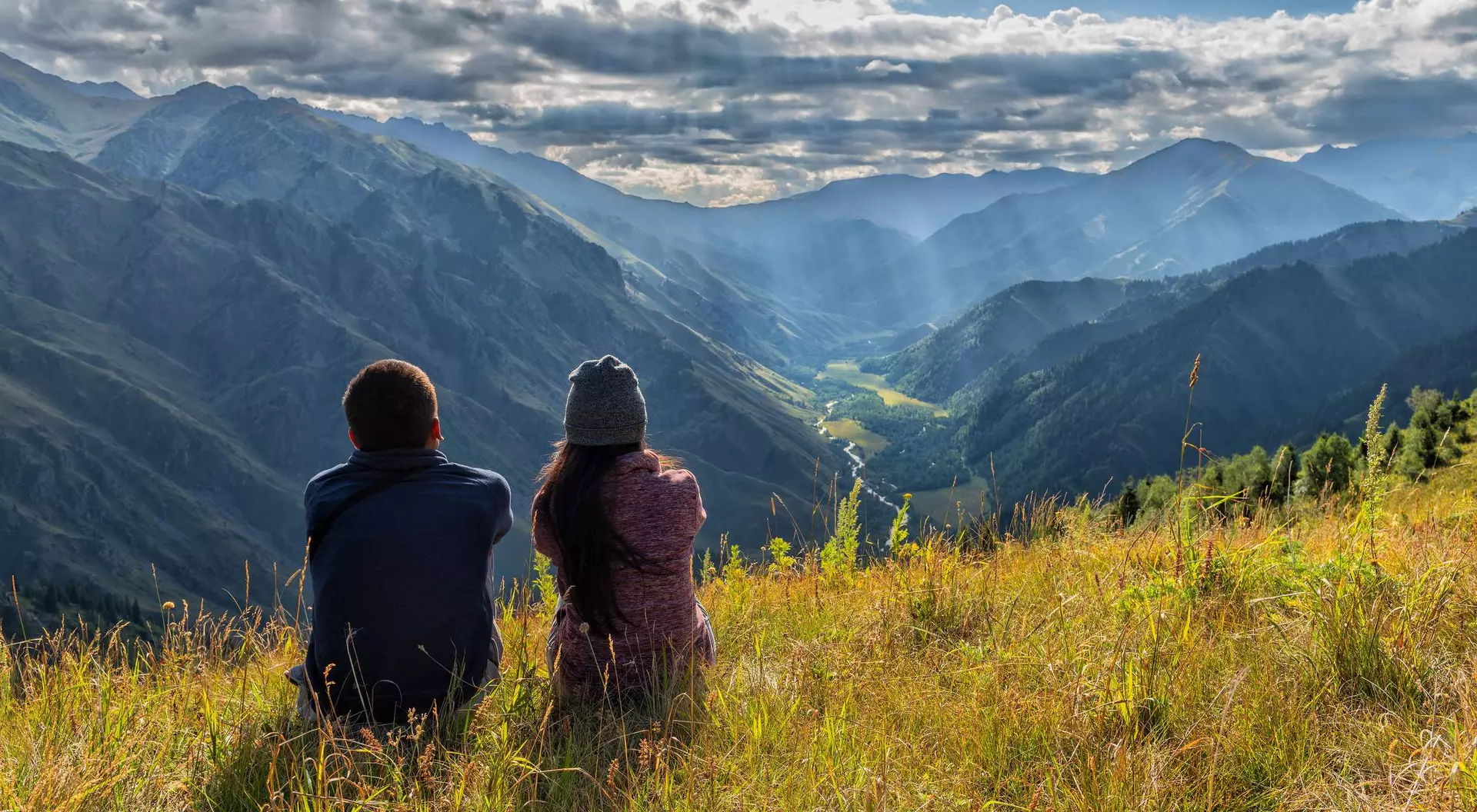 Two people sit on a grassy hillside, gazing at a panoramic view of mountains and valleys under a cloudy sky.
