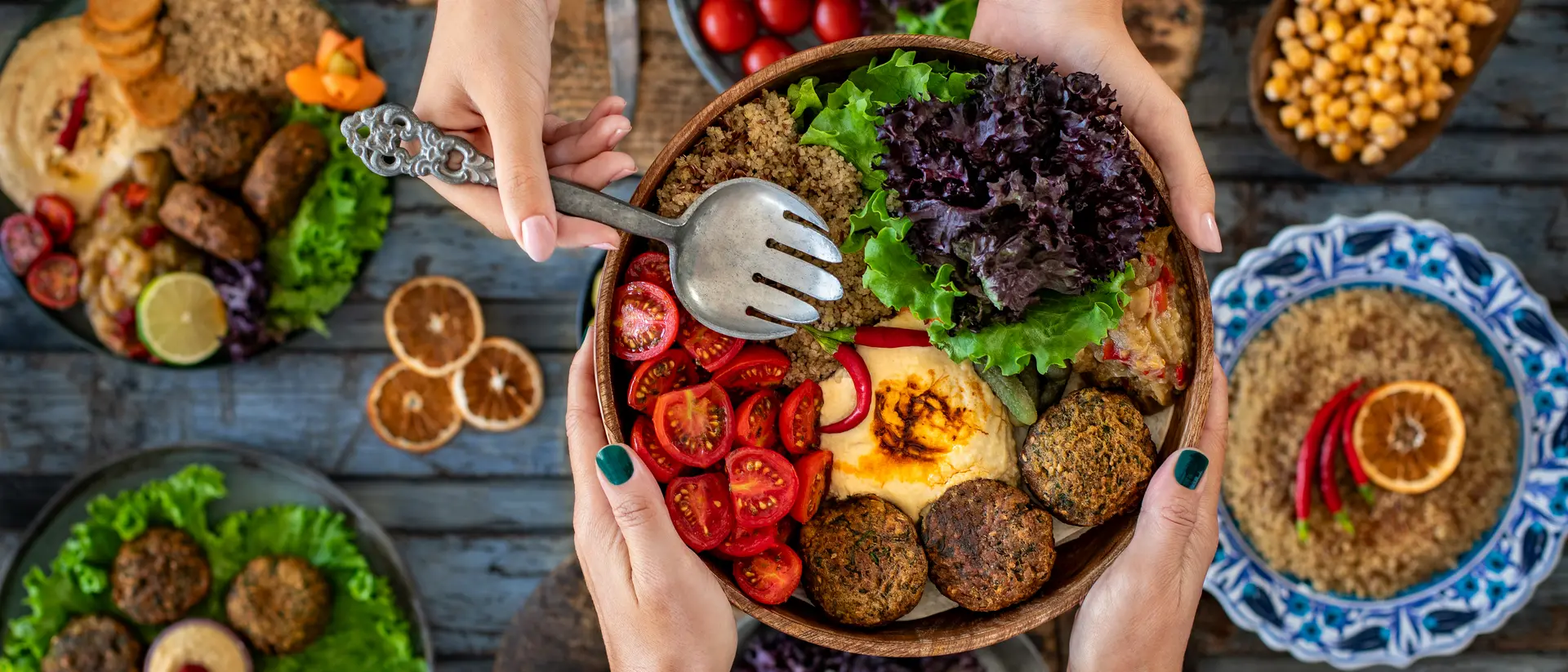 A colorful spread of salads, falafel, and vegetables, with hands reaching for a bowl on a rustic wooden table.