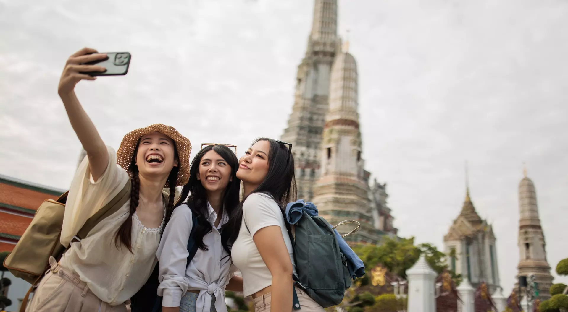 Three smiling women take a selfie in front of a temple with intricate architecture on a cloudy day.