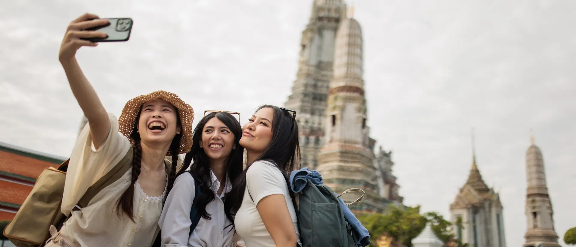 Three smiling women take a selfie in front of a temple with intricate architecture on a cloudy day.