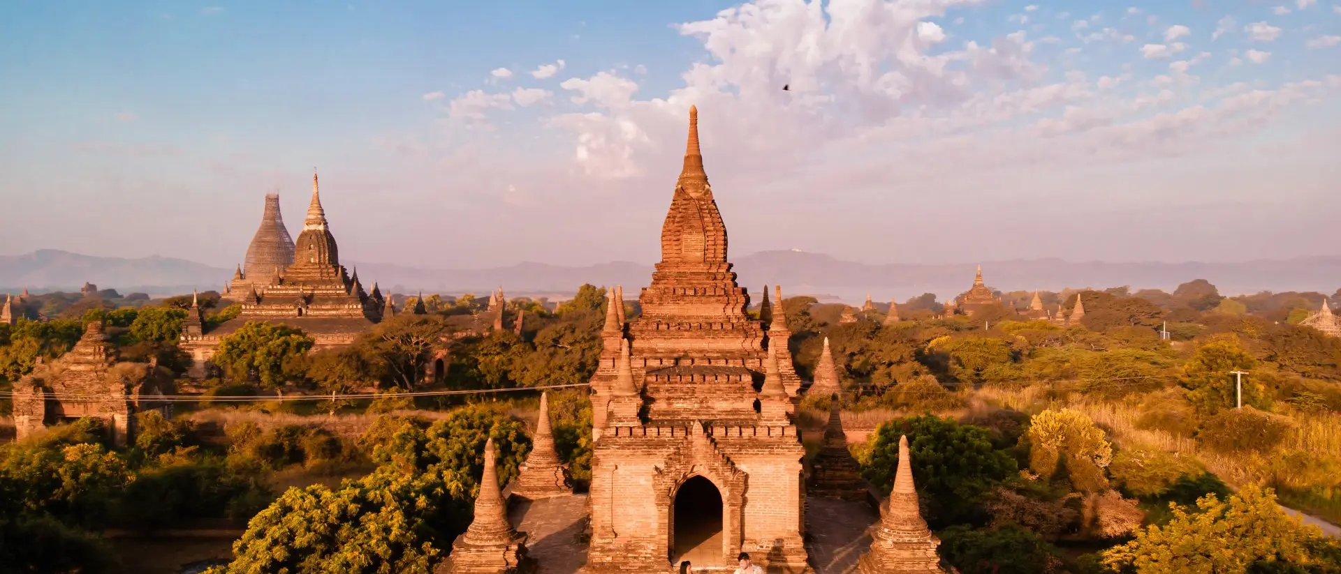 Aerial view of ancient temples in Bagan, Myanmar, with two people exploring among greenery under a blue sky.