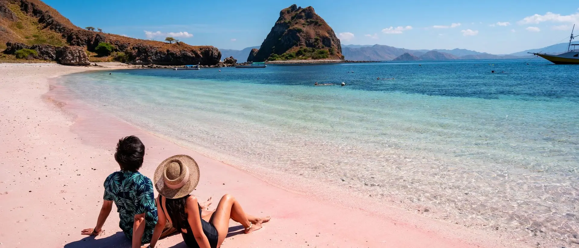 Two people relax on a pink beach, looking out at clear blue water and a rocky island in the distance under a sunny sky.
