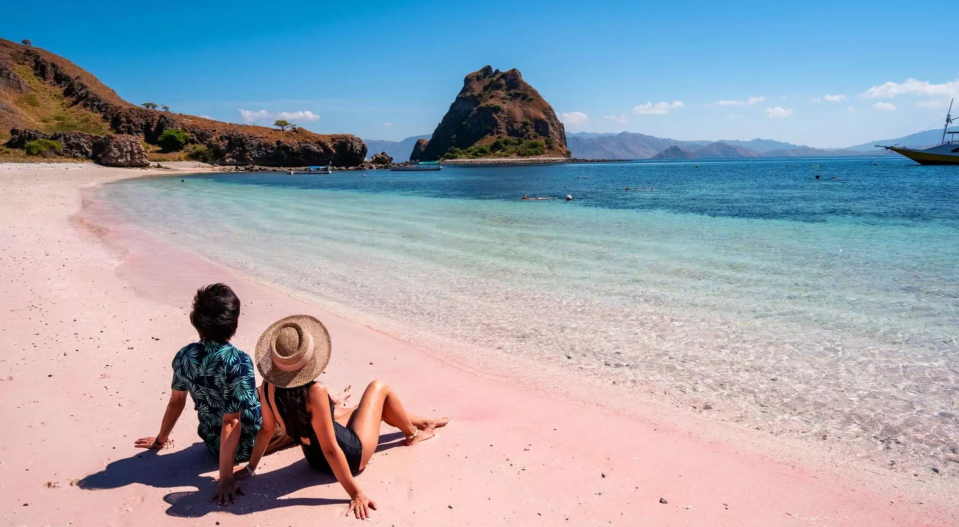 Two people relax on a pink beach, looking out at clear blue water and a rocky island in the distance under a sunny sky.