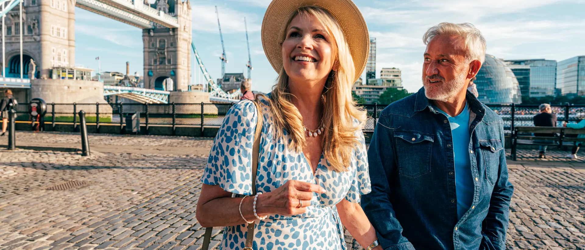 A smiling couple strolls hand in hand near the Tower Bridge in London on a sunny day.