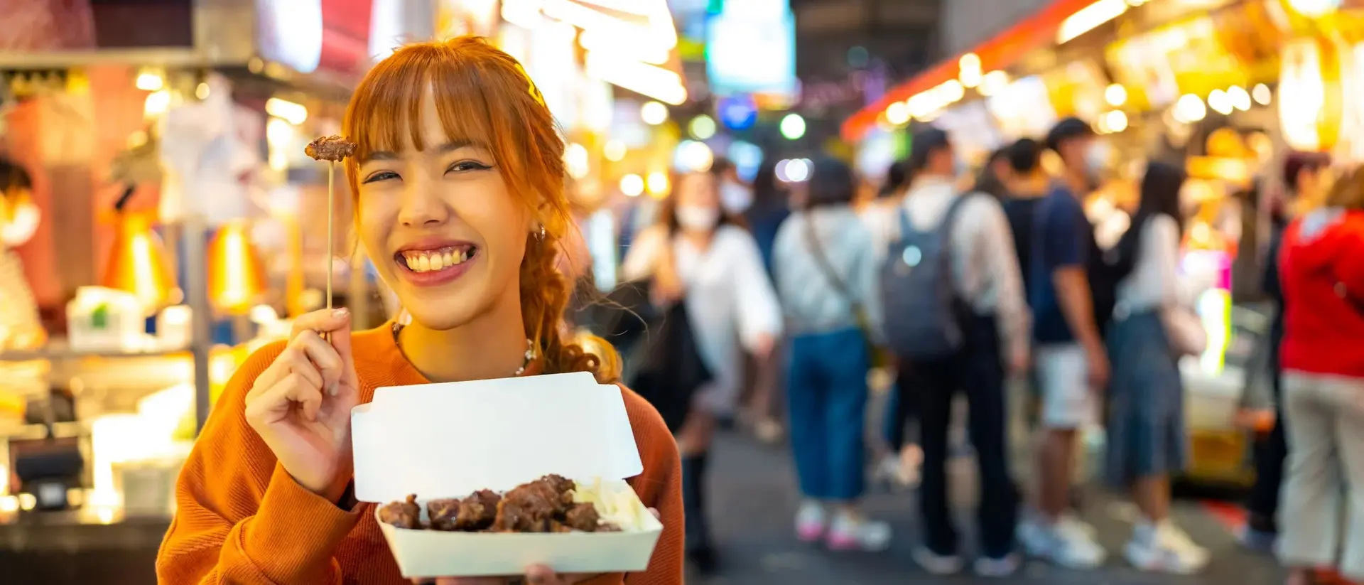 A smiling woman holds a takeout box of food at a busy night market filled with colorful stalls and crowds.