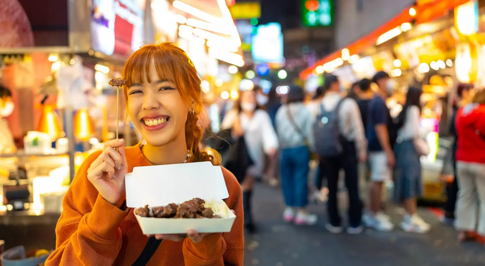 A smiling woman holds a takeout box of food at a busy night market filled with colorful stalls and crowds.