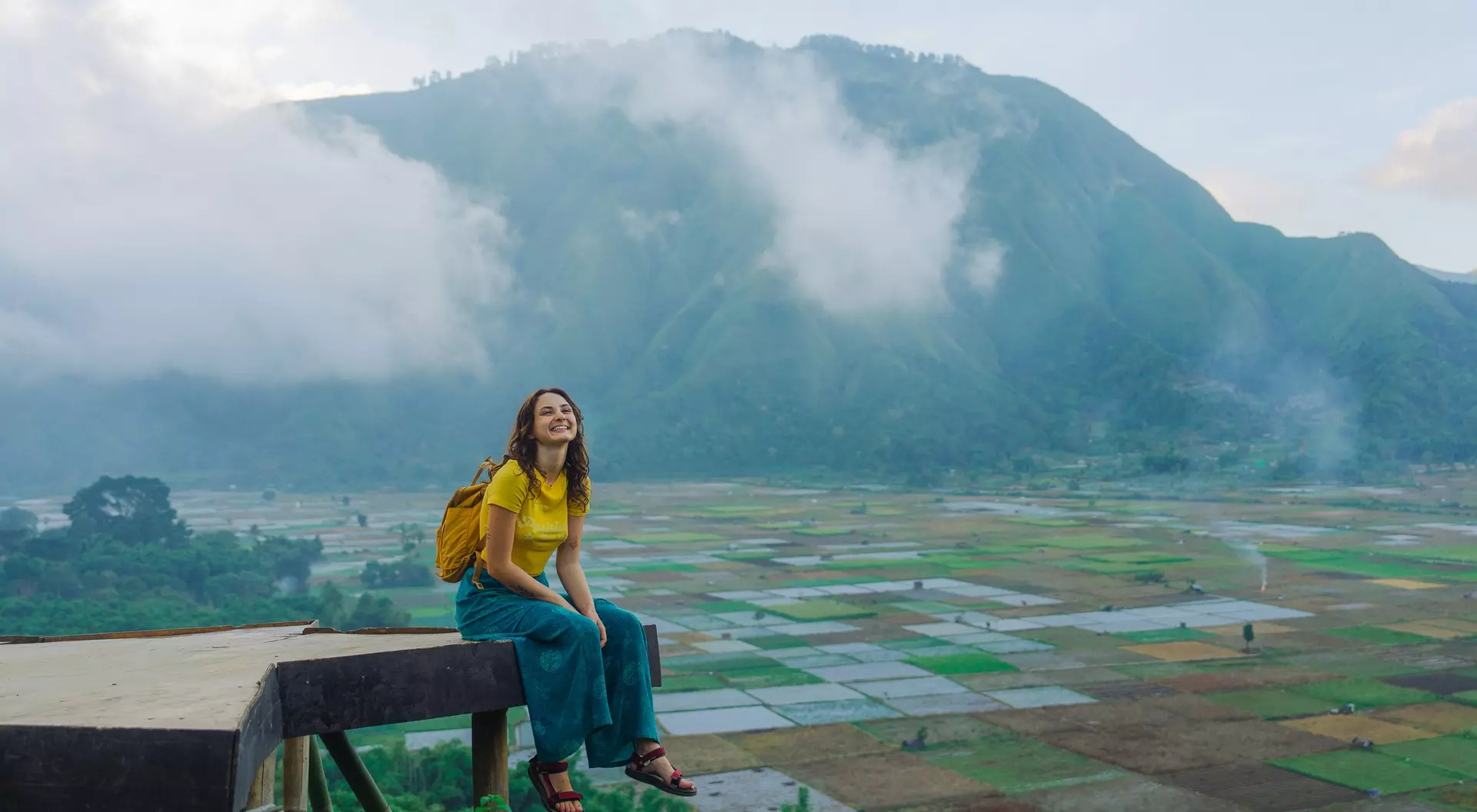 A smiling person in yellow sits on a ledge, surrounded by lush fields and mountains shrouded in clouds.