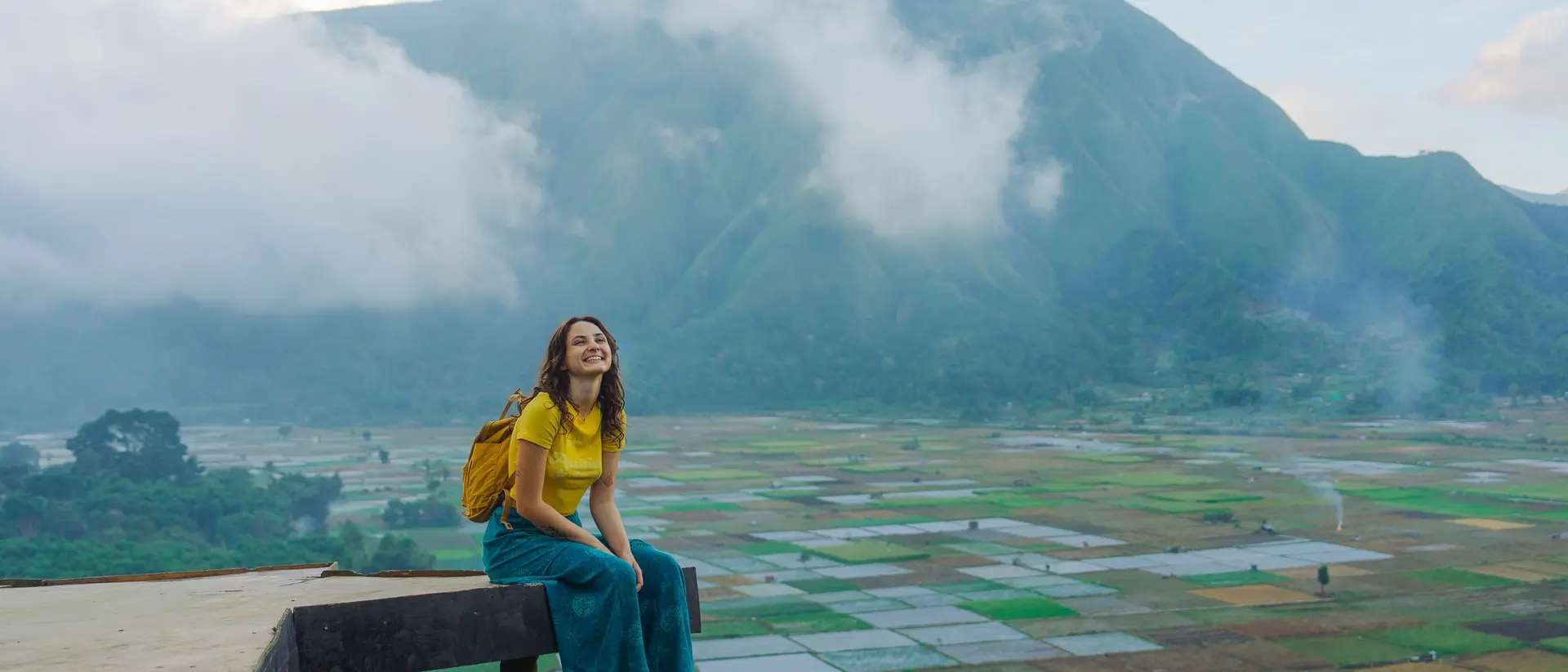 A smiling person in yellow sits on a ledge, surrounded by lush fields and mountains shrouded in clouds.