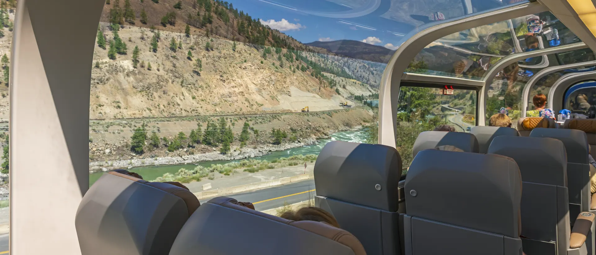Interior view of a scenic train car, with plush seats and large windows showcasing mountains and a river outside.