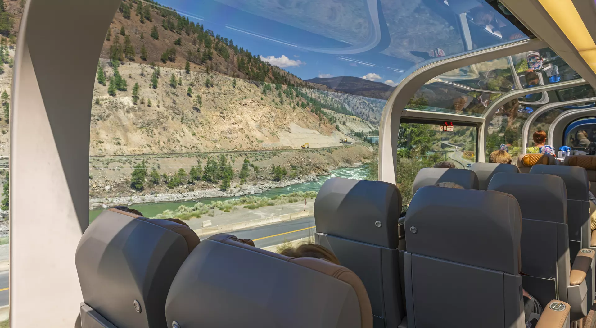 Interior view of a scenic train car, with plush seats and large windows showcasing mountains and a river outside.